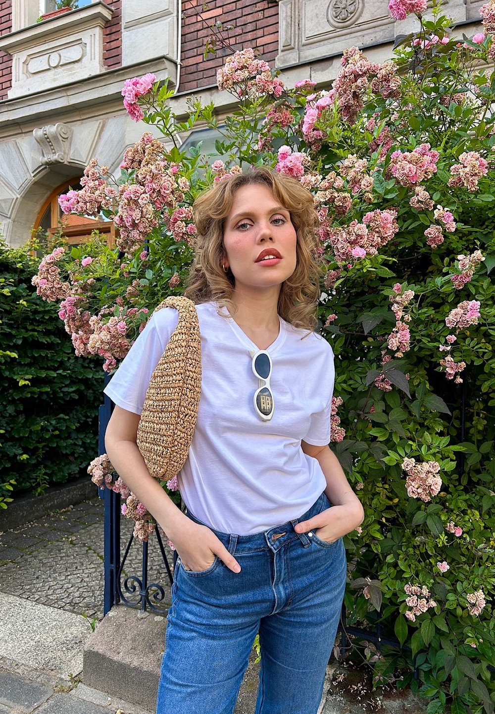 T-shirt blanc, jeans en denim bleu et un sac beige tissé. Lunettes de soleil rondes avec un détail en argent. Des roses roses en fleurs en arrière-plan.