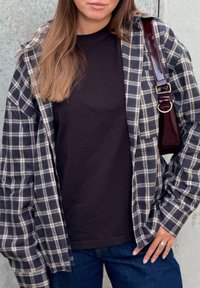 Nagy méretű kockás flanel ing tengerészkék és bézs színekben, fekete pólóra rétegezve. Csillogó bordó táskapánt és kék farmer mellé.