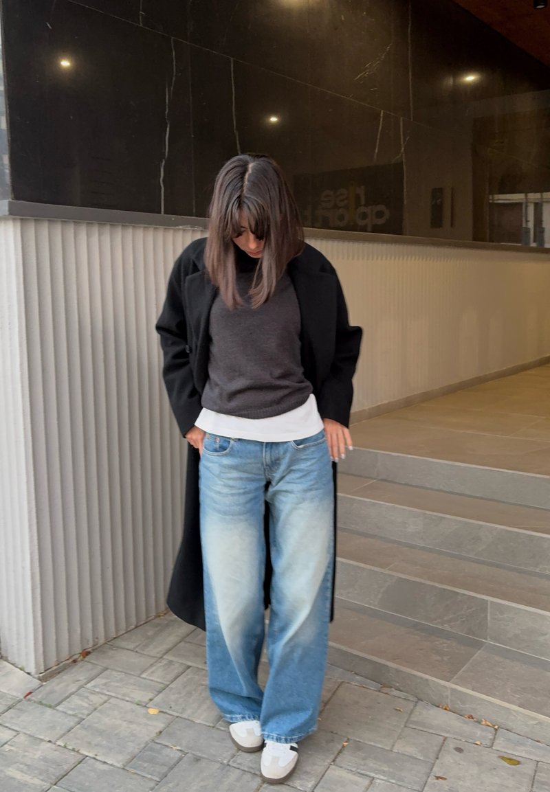 Femme portant un manteau noir, un pull foncé, une chemise blanche, un jean large bleu et des baskets blanches, debout près d'escaliers et d'un mur texturé.