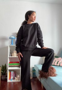 Vrouw in zwart sweatshirt en broek met harige slippers poseert met éénBeen op een blauwe bank in een gezellige woonkamer.