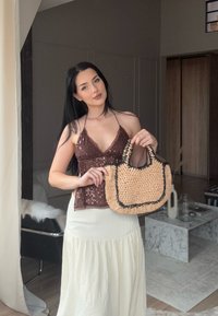Donna con lunghi capelli scuri che indossa un top halter marrone con paillettes e una gonna bianca tiene in mano una borsa di paglia intrecciata in un soggiorno moderno.