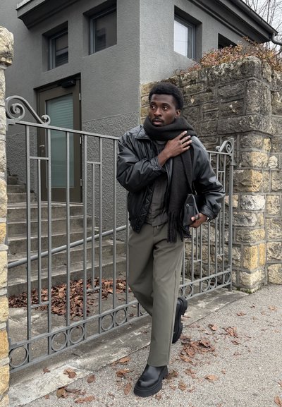 Hombre con chaqueta de cuero negra, bufanda negra, pantalones grises y botas negras caminando cerca de un muro de piedra y una puerta metálica fuera de un edificio.