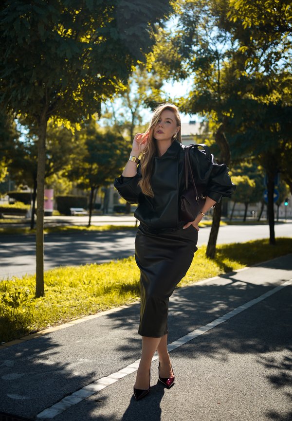 Mujer con chaqueta de cuero negra y falda con tacones altos está en una acera flanqueada por árboles bajo la luz del sol, sosteniendo un bolso al hombro.