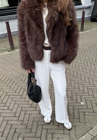 Giacca in pelliccia sintetica marrone, top bianco, pantaloni bianchi, borsa intrecciata nera. Sneakers grigie con lacci bianchi. Design dell'outfit testurizzato e a strati.