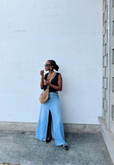 Top negro sin mangas, falda de mezclilla clara de tiro alto con una abertura frontal, zapatos de tacón negros y un bolso cruzado de mimbre.