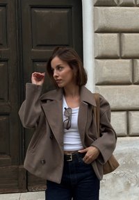 Hnědá oversized bunda ze směsi vlny s velkými klopami, černými knoflíky, nošená přes bílý přiléhavý top a tmavé džíny. Jednoduchá taška na řemínku.