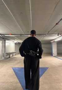 Čierny sveter s textúrou a široké čierne nohavice doplnené malou čiernou kabelkou cez rameno, v slabozosvetlenej parkovacej garáži.