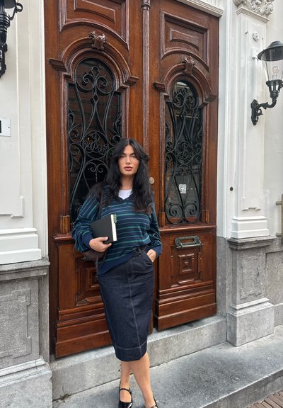 Suéter a rayas en navy con detalles en verde, combinado con una falda lápiz de mezclilla oscura. Sosteniendo un cuaderno, de pie junto a unas puertas de madera ornamentadas.