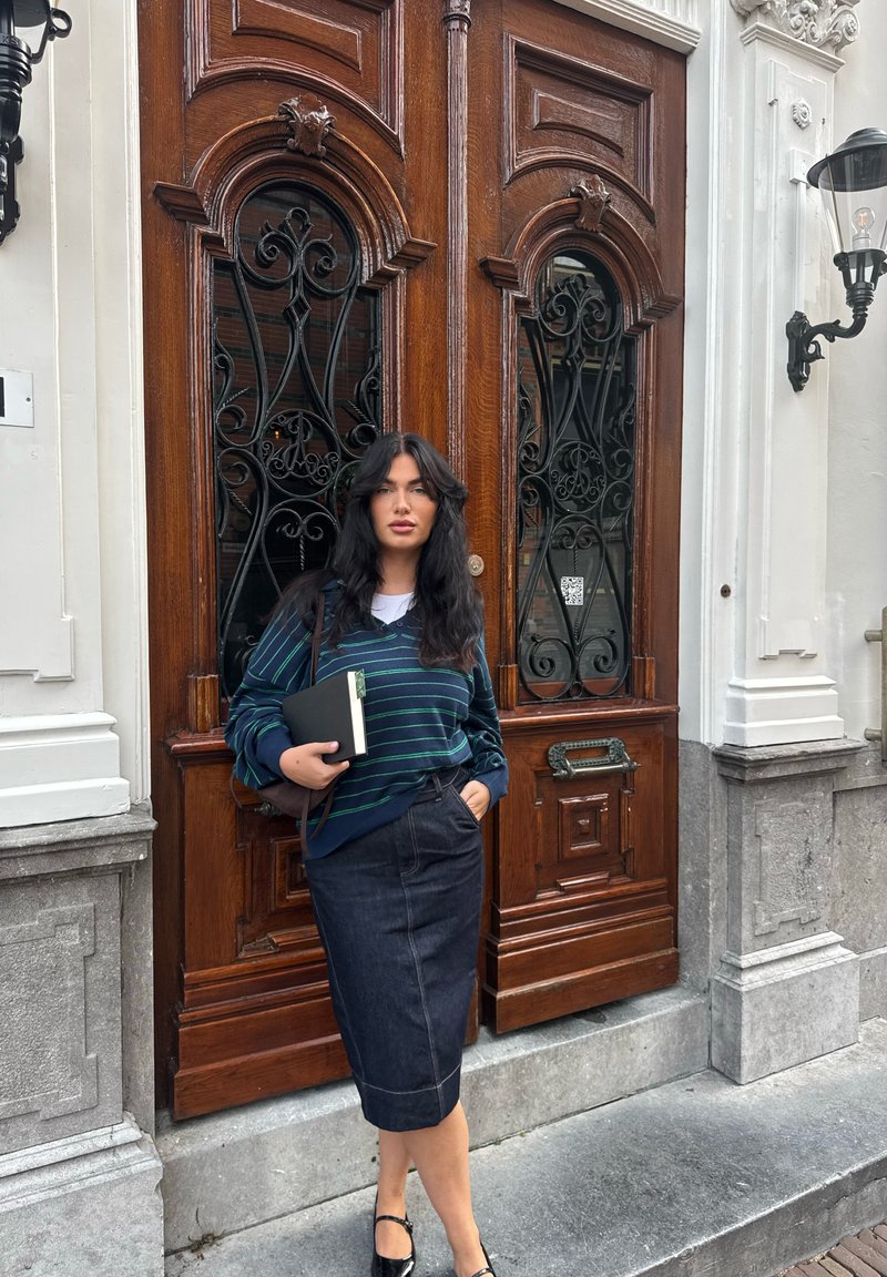 Suéter listrado azul-marinho com detalhes verdes, combinado com uma saia lápis de jeans escuro. A segurar um bloco de notas, em pé ao lado de portas de madeira ornamentadas.