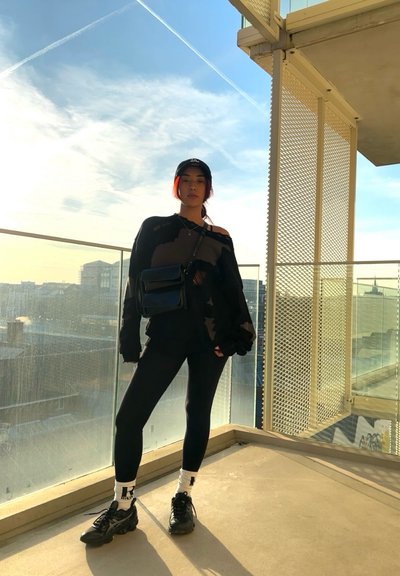 Suéter negro oversized con detalles desgastados, leggings negros y zapatillas deportivas negras. Accesorios incluyen un pequeño bolso negro cruzado y una gorra.