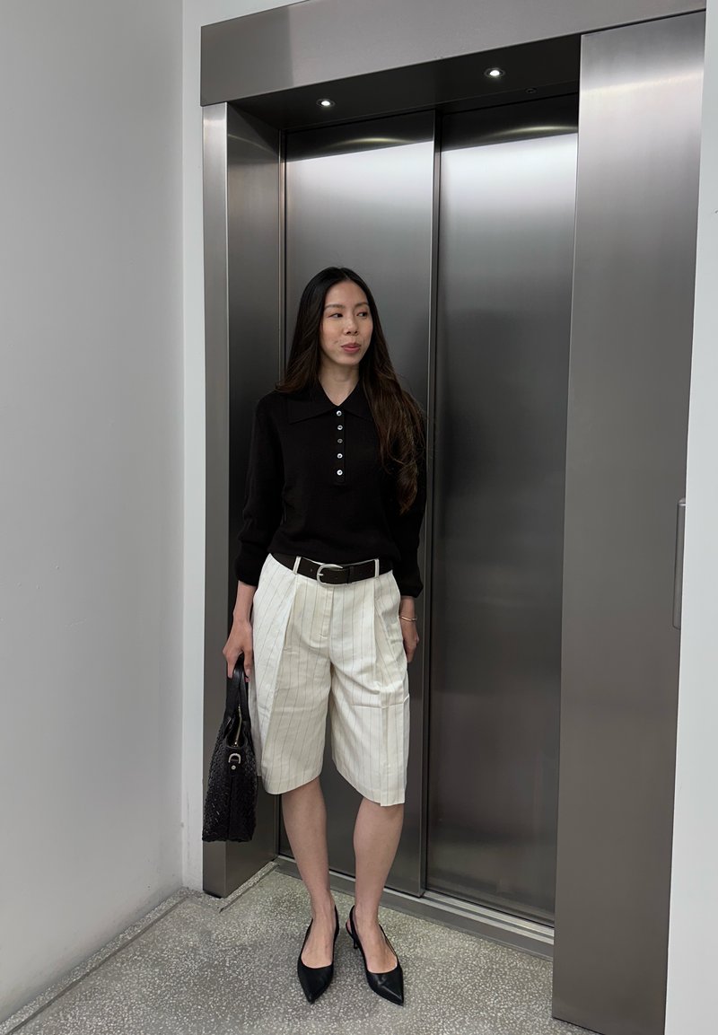 Donna in piedi davanti a porte chiuse di ascensore in acciaio inox, indossa un top nero, pantaloncini bianchi a righe sottili e tacchi neri, tiene una borsa nera.