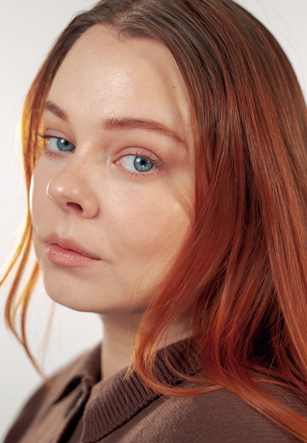 Jeune femme aux longs cheveux roux et aux yeux bleus, portant un pull marron, regardant légèrement par-dessus son épaule sur un fond clair uni.