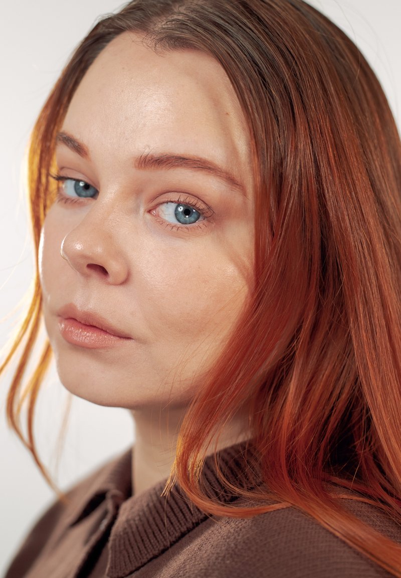 Jeune femme aux longs cheveux roux et aux yeux bleus portant un pull marron, regardant légèrement par-dessus son épaule sur un fond clair uni.