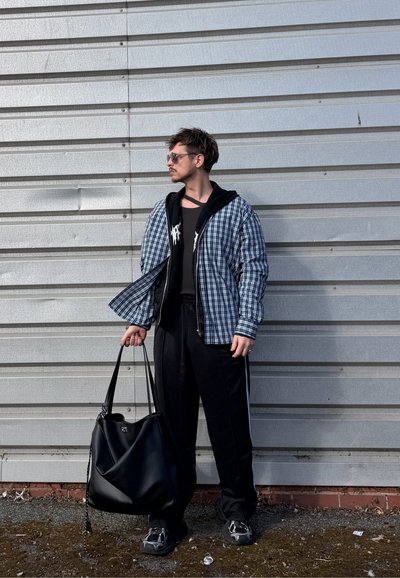 Hombre con gafas de sol está junto a una pared metálica, lleva chaqueta de cuadros, pantalón negro y zapatillas, sostiene una gran bolsa negra y mira hacia la izquierda.