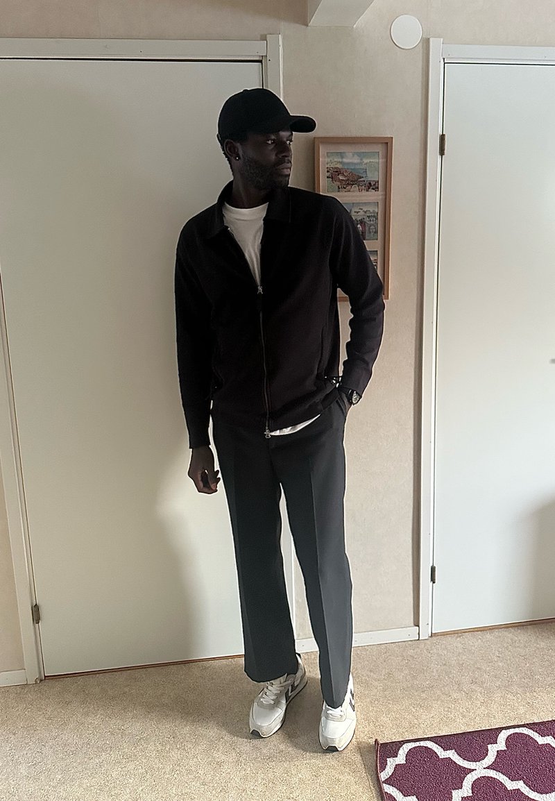 Άνδρας με μαύρο καπέλο, μαύρο μπουφάν, λευκό πουκάμισο, σκούρο παντελόνι και λευκά αθλητικά παπούτσια στέκεται σε εσωτερικό χώρο κοντά σε κλειστές λευκές πόρτες και μια κορνιζαρισμένη εικόνα.