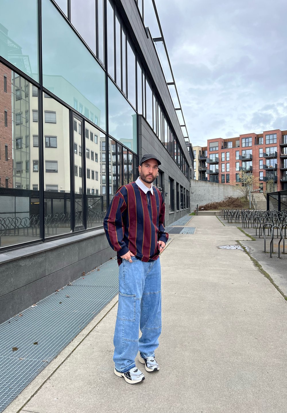 Maglione a righe in blu navy e bordeaux. Pantaloni cargo leggermente blu. Cappellino nero e sneakers con dettagli bianchi. Sfondo urbano con finestre di vetro.