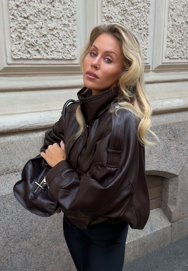 Femme aux longs cheveux blonds portant une veste en cuir marron foncé et tenant un sac à main en cuir assorti devant un mur en pierre texturé.