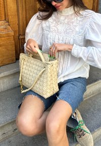 Beige vævet handbag med en struktureret form, der har et flet håndtag og gyldne hardware-detaljer. Modellen har en hvid bluse og denim shorts på.