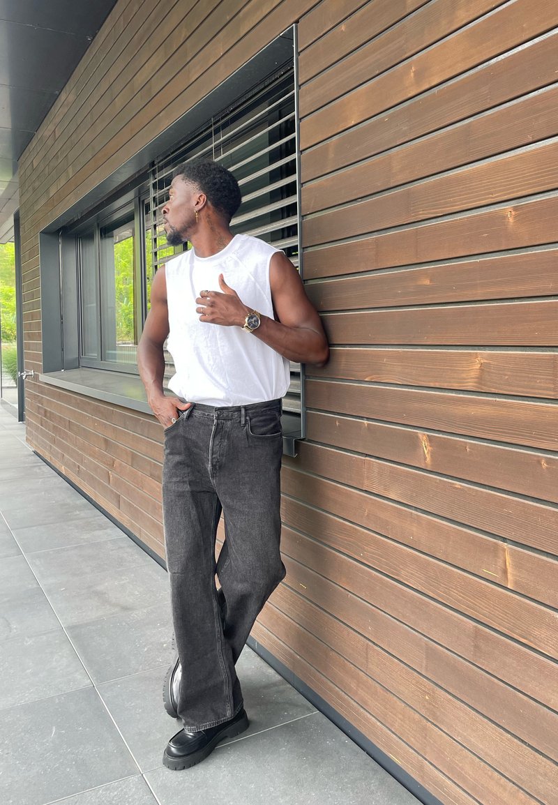 Chemise sans manches blanche, jean large gris foncé et chaussures noires épaisses, s'appuyant contre un mur en bois avec des lattes horizontales.