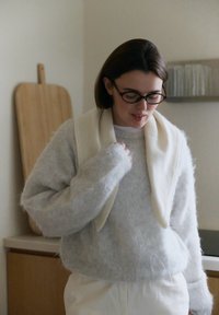 Pull gris clair duveteux avec larges poignets, superposé sur un haut blanc. Une écharpe blanche drapée autour du cou. Planche à découper en bois naturel en arrière-plan.