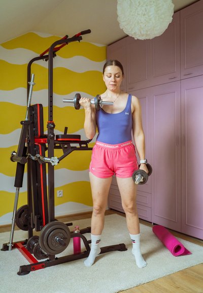 Mujer con ropa de entrenamiento levantando pesas en el interior, junto a una máquina de pesas, una esterilla de yoga y un fondo de pared ondulada y colorida.