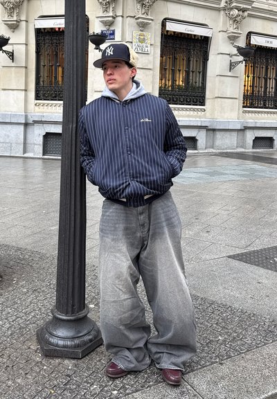 Persona con una chaqueta de raya diplomática azul marino sobre una sudadera gris, usando jeans anchos grises y zapatos burdeos, apoyándose en una farola.