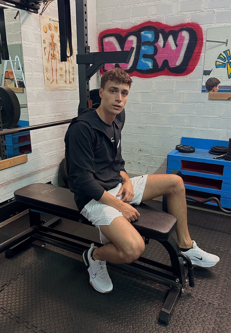 Jeune homme en veste noire et short blanc assis sur un banc de gym près d'équipements de musculation et de graffitis sur un mur en briques blanches.
