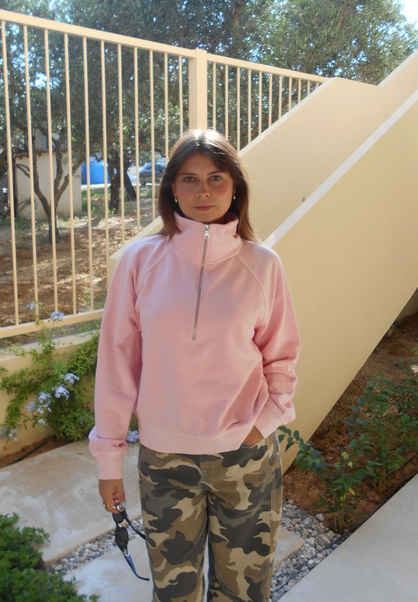 Junge Frau steht im Freien nahe einer Treppe und einem Geländer, trägt ein hellrosa Reißverschlussoberteil und Tarnhosen, hält eine Sonnenbrille in einer Hand.