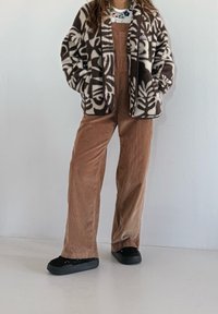 Salopette en velours côtelé marron à jambes larges, associée à une veste moelleuse à motifs marron et crème. Des chaussures noires complètent la tenue.