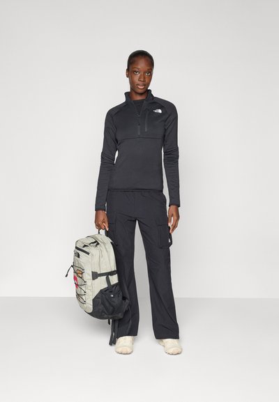 Suéter negro de manga larga con cuello de cremallera, pantalones cargo negros y una mochila bicolor con bolsillo de malla y detalles de logo.