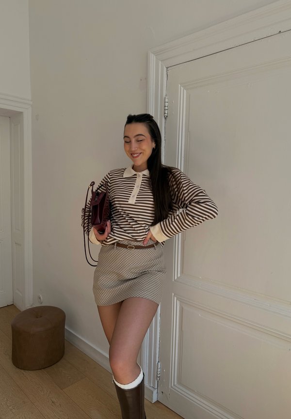 Femeie în cămașă cu mânecă lungă în dungi, fustă mini cu model și cizme maro, ținând o geantă burgundy, stând în interior lângă o ușă albă.
