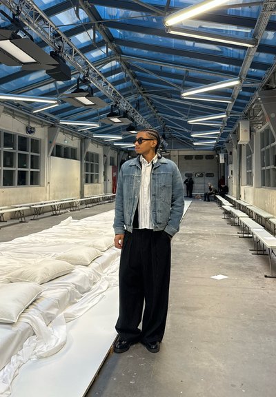 Chaqueta de mezclilla sobre una camisa blanca, combinada con pantalones anchos de rayas negras y zapatos negros, ambientada en un entorno industrial espacioso.