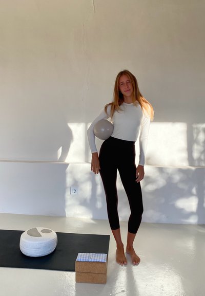 Mujer con una blusa blanca de manga larga y leggings negros, sosteniendo una bola gris. Esterilla de yoga negra con un cojín de yoga blanco y un bloque de corcho cerca.