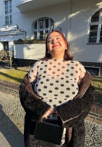 Manteau noir duveteux porté sur un haut gris transparent à pois noirs. Tient un petit sac à main noir embossé effet croco. La lumière du soleil projette des ombres sur la scène.