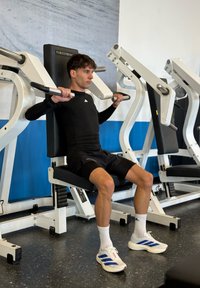 Ülő súlyzógép fekete háttámlával, állítható karfogantyúkkal, és egy fekete hosszú ujjú felsőt viselő felhasználóval, aki gyakorlatot végez.