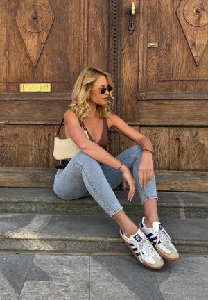 Mujer con cabello rubio y gafas de sol sentada en unos escalones de piedra frente a una gran puerta de madera, vestida con vaqueros, una camiseta marrón y zapatillas blancas.