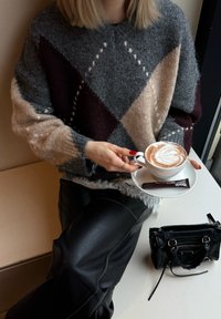 Hall ruuduline halli, burgundi ja beeži värvitooniga kampsun, millel on liiga suur suurus. Hoides käes valget tassi, mille peal on lattekunstiga latte, ümmargusel taldrikul ja musta kotti kõrval.