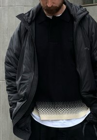 Sort boblejakke over en svart poloshorte med en teksturert kremfarget kant med et prikkete mønster; hvit skjorte under.