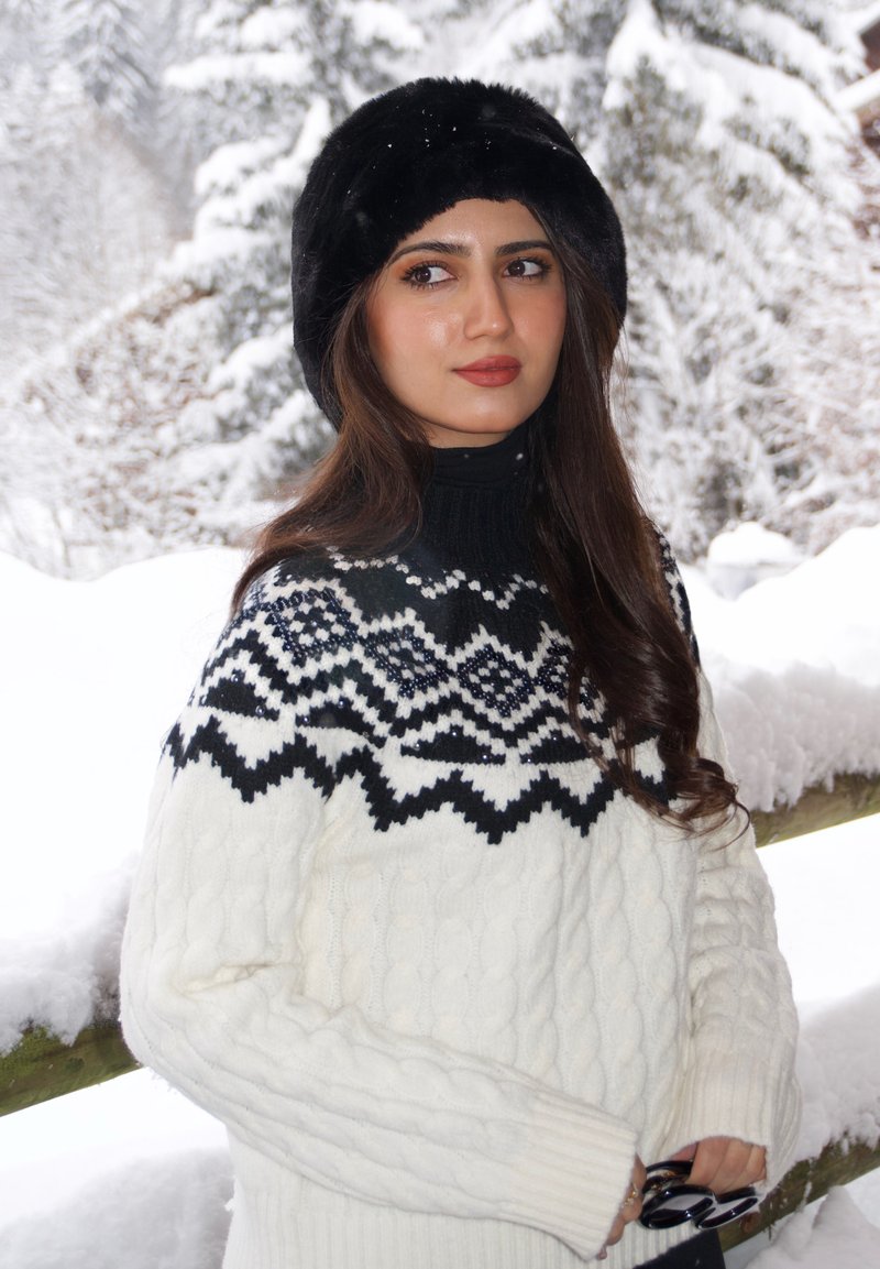 Pull en laine blanc à torsades avec un empiècement noir et blanc à motifs, associé à un bonnet en fourrure noir. Elle tient des lunettes de soleil dans sa main.