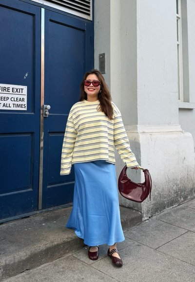 Mujer con top amarillo a rayas y falda azul está sonriendo junto a una puerta azul, sosteniendo un bolso granate y llevando zapatos a juego.