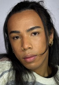 Close-up van een persoon met middelbruin huid, lang zwart haar naar achteren gebonden, met een zilveren hoepeloorbel en een neusring, en zachte roze lippen.