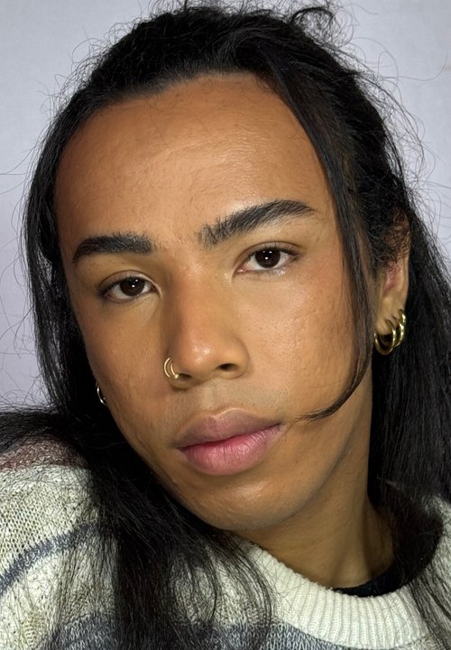 Primo piano di una persona con pelle marrone media, capelli neri lunghi raccolti, che indossa un orecchino a cerchio d'argento e un anello al naso, con labbra rosa tenue.