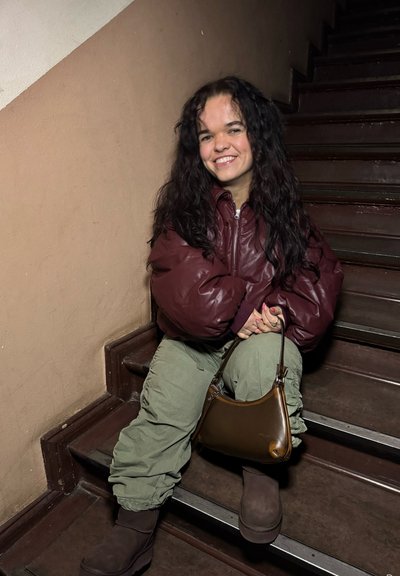 Chaqueta acolchada marrón, pantalones cargo verdes oliva, bolso marrón y botas marrones oscuras. Sentada en escaleras de madera con fondo de pared texturizada.