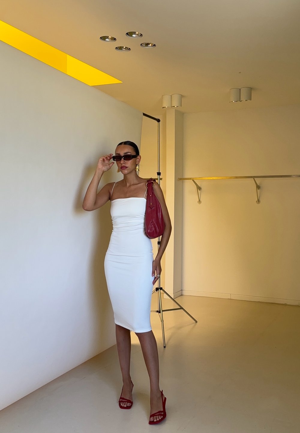 Abito bodycon bianco con spalline sottili, borsa rossa, sandali con tacco rosso e occhiali da sole. Sfondo minimalista con illuminazione soffusa.