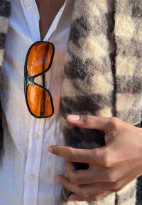 Orange sunglasses dangle from a light-coloured, button-up shirt, layered with a brown and beige patterned fabric. Smooth textures are apparent.