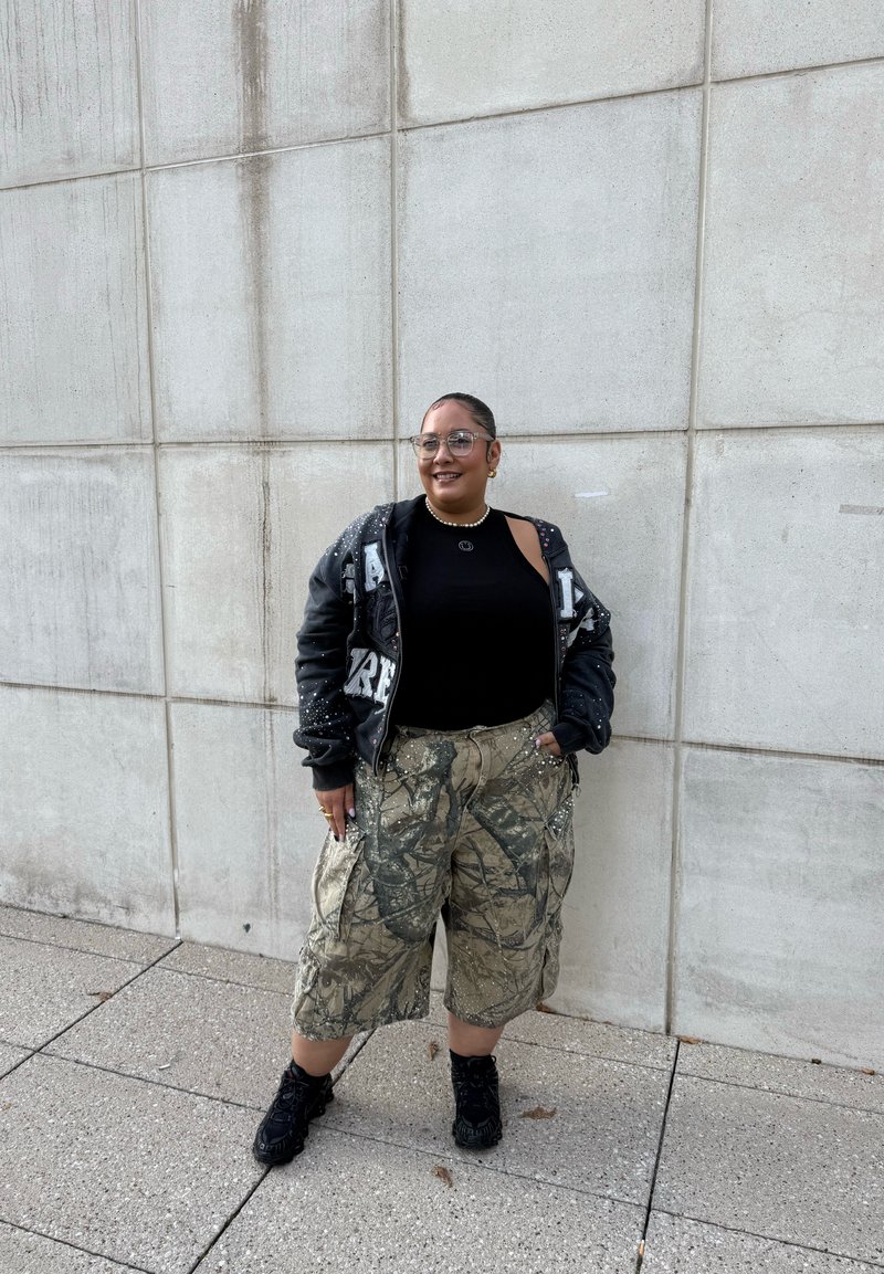 Black graphic hoodie, black tank top, camo cargo shorts, black sneakers. Accessories include a pearl necklace and glasses. Standing against a wall.