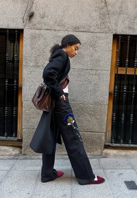 Zwarte wijde broek met kleurrijke floral borduursels, gecombineerd met een donkere jas en bordeaux slip-ons. Bruine gestructureerde handtas.