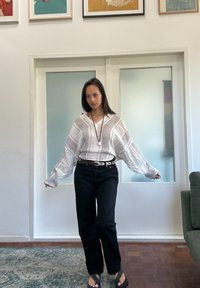 Svetlo šedá pruhovaná oversized blúzka s dlhými rukávmi, kombinovaná s čiernymi nohavicami s vysokým pásom a čiernymi sandálmi. Viditeľná drevená podlaha a sklenené dvere.