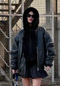Črna usnjena bomber jakna s kapuco iz umetnega krzna, oblečena čez črno obleko z nabrano obrobo, dopolnjena z angularnimi sončnimi očali in torbico.