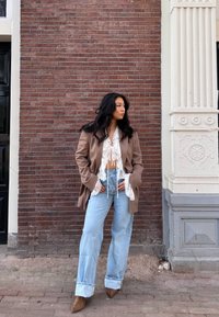 Brun oversized blazer, hvid blonde top, lys blå vidde jeans med opbrettede ben, og brune spidse sko mod en murstensvæg.